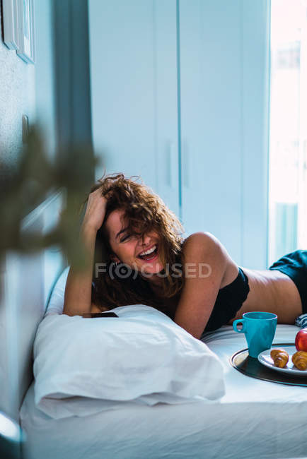 Laughing woman lying on bed with breakfast and looking at camera — Stock Photo
