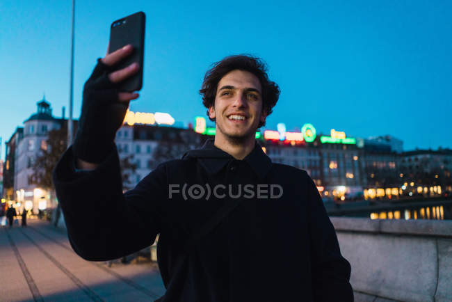 Улыбающийся мужчина в теплой одежде стоит на вечерней улице и делает селфи со смартфоном . — стоковое фото