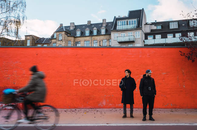Two men in warm clothes standing by orange wall on street scene. — Stock Photo