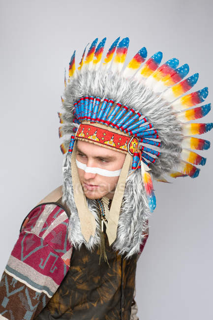 Man with white line on face posing in traditional Native American costume in studio and looking aside — Stock Photo