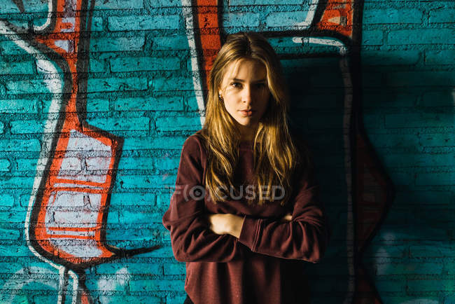 Jolie jeune femme avec les bras croisés posant au mur de briques avec graffiti et regardant la caméra — Photo de stock