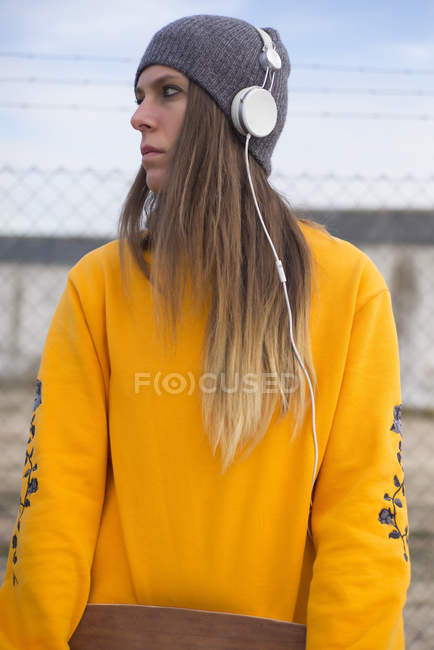 Young girl in headphones posing with longboard in hands and looking away at street scene — Stock Photo