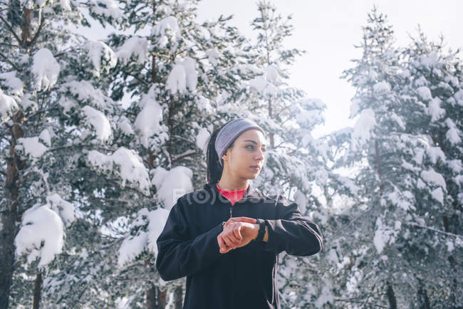 Спортивная женщина держит за руку спортивные часы в зимнем лесу и смотрит в сторону — стоковое фото