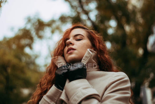 Молодая рыжая женщина с закрытыми глазами регулирует воротник пальто — стоковое фото