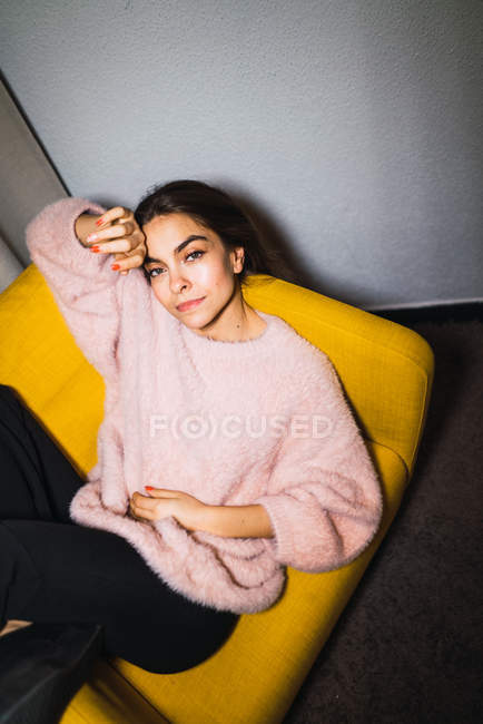 Mujer joven en suéter rosa relajándose en sillón en casa y mirando a la cámara - foto de stock