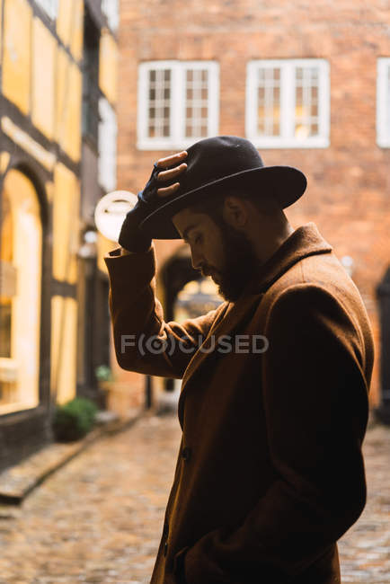 Side view of bearded man wearing coat putting on hat at street scene — Stock Photo