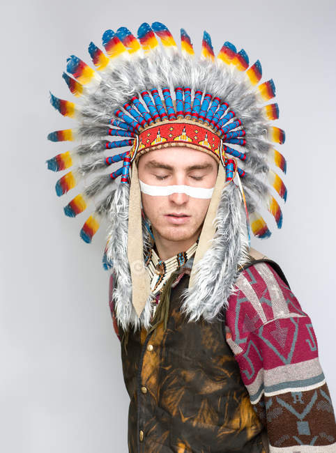 Portrait of man with line on face posing in Native American costume with eyes closed on white background — Stock Photo
