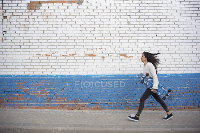 Vista laterale della ragazza bruna che cammina con longboard lungo il muro della strada — Foto stock