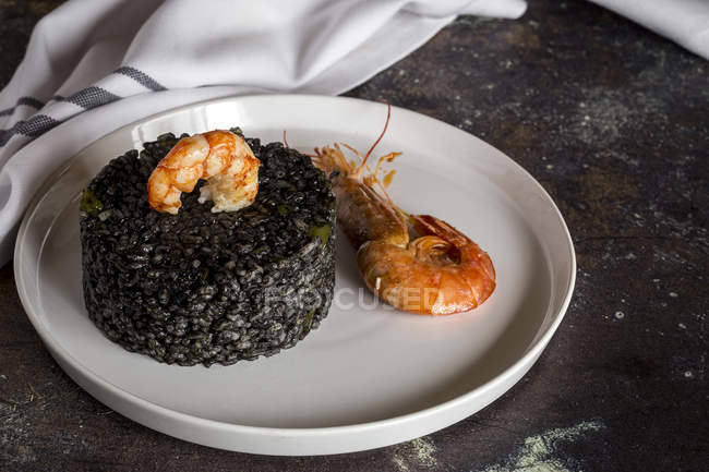 Still life of black rice with prawns on white plate on old table. — Stock Photo