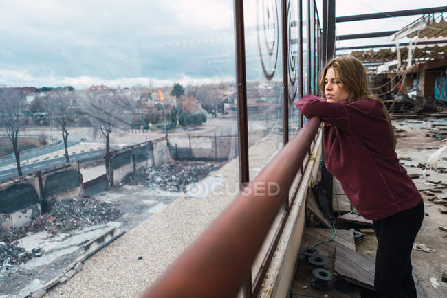 Side view of thoughtful young woman leaning on handrail and looking away at window of grungy building. — Stock Photo
