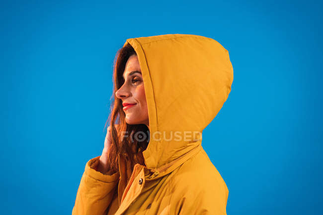 Mulher sorridente no capuz amarelo posando contra fundo azul — Fotografia de Stock