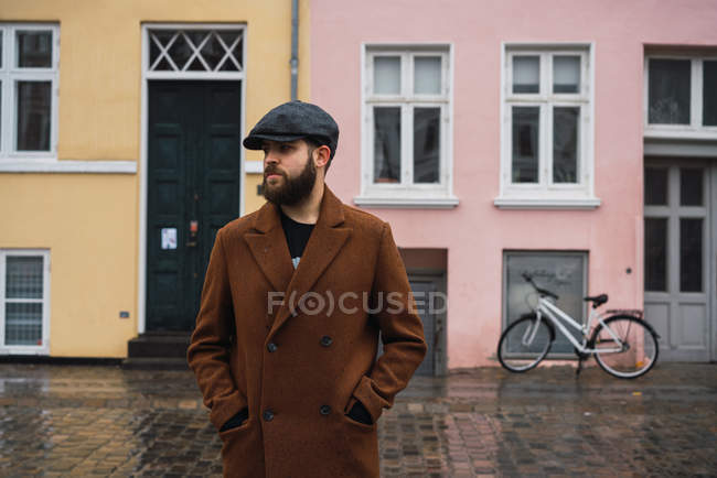 Bärtiger Mann im Vintage-Mantel posiert vor dem Hintergrund eines an Gebäuden geparkten Fahrrads — Stockfoto