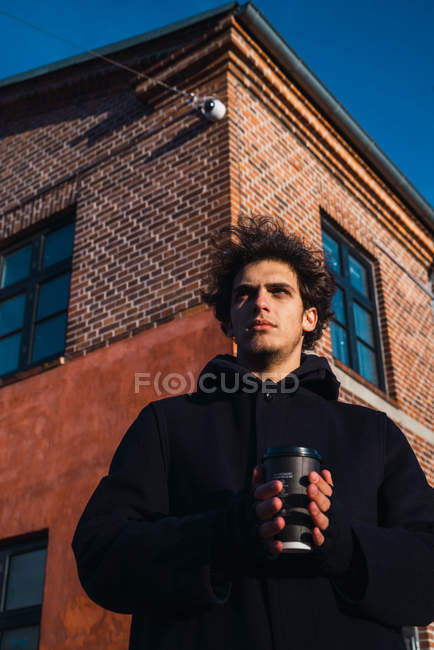 Молодий чоловік у теплому одязі стоїть з чашкою кави і дивиться на вулицю . — стокове фото