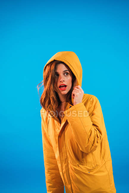 Femme expressive en capuche jaune posant sur fond bleu — Photo de stock