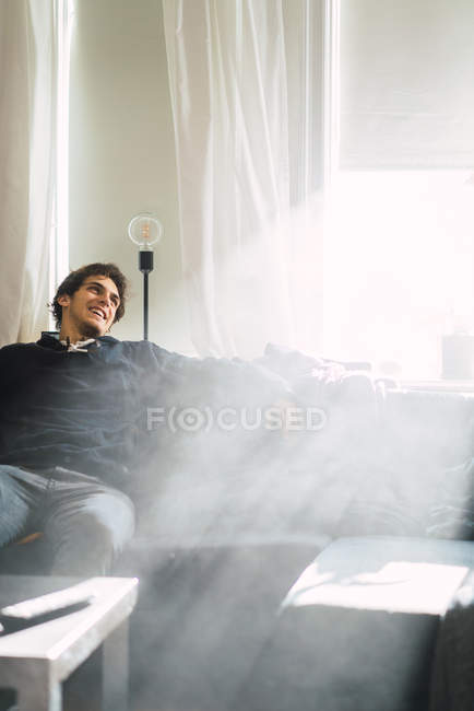 Cheerful man sitting coach in smoky or steamy room at home. — Stock Photo
