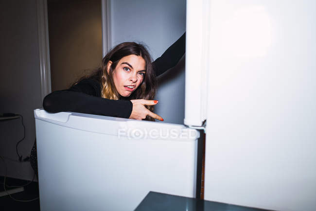 Mulher expressiva repugnante olhando para a câmera e apontando para a geladeira aberta . — Fotografia de Stock