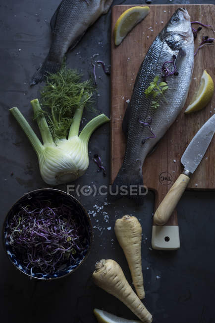 Bodegón de pescado crudo sobre tabla de madera y verduras sobre mesa - foto de stock