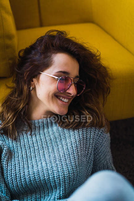 Mulher sorridente de óculos sentada no chão e apoiada na poltrona — Fotografia de Stock