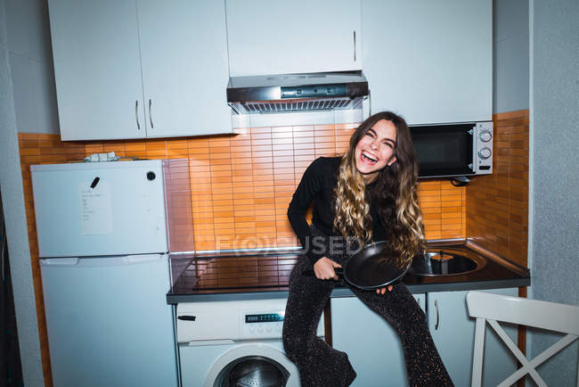 Rindo mulher sentada na mesa da cozinha com panela nas mãos — Fotografia de Stock
