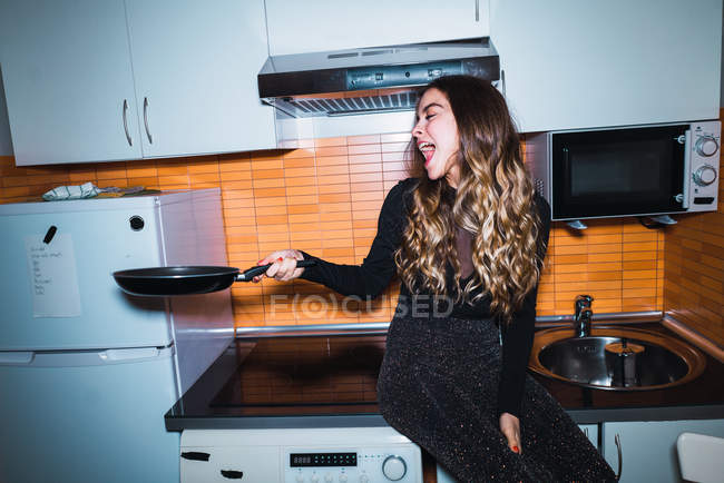 Mujer riendo sentada en la mesa de la cocina y extendiendo la mano con sartén - foto de stock