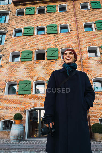 Веселий молодий фотограф стоїть на цегляній будівлі на вулиці і дивиться геть — стокове фото