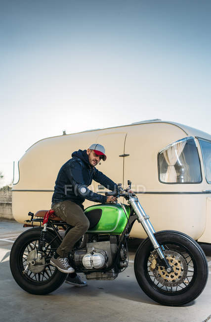 Портрет стильного мужчины, сидящего на мотоцикле возле фургона и смотрящего в камеру — стоковое фото