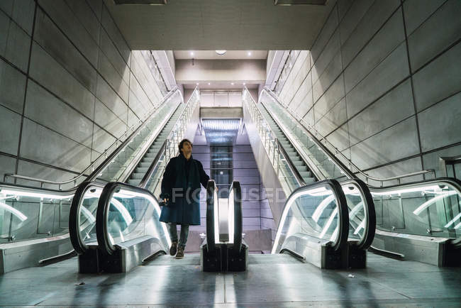 Young man in coat walking up on moving stairs and looking up. — Stock Photo