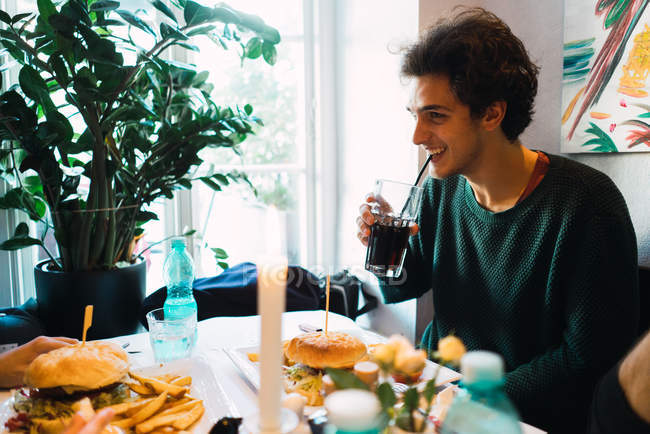 Homem alegre bebendo coca e comendo hambúrguer na mesa do café — Fotografia de Stock