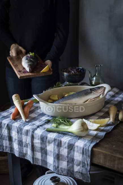 Crop person carrying fish on wooden board by table with ingredients — Stock Photo