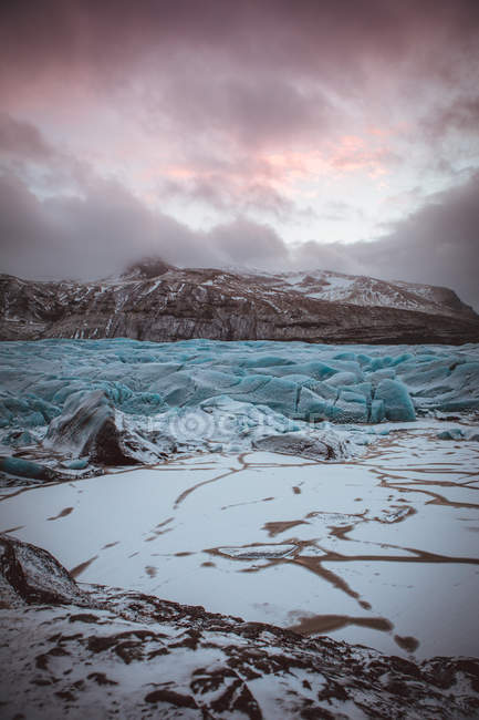 View of frost glacier surface with background of rocky cliffs in gloomy clouds. — Stock Photo