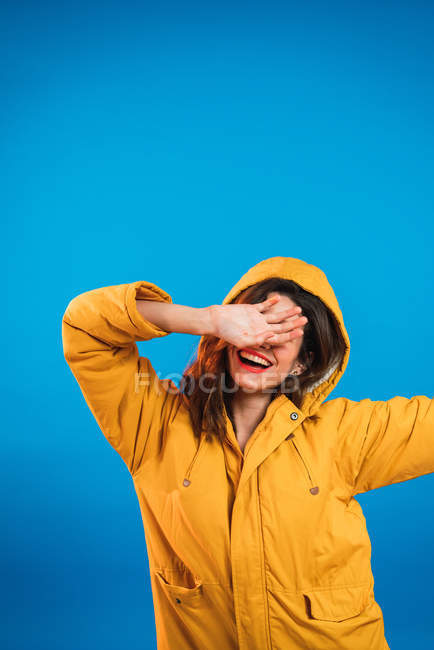 Улыбающаяся молодая женщина в жёлтой куртке закрывает лицо рукой на синем фоне . — стоковое фото