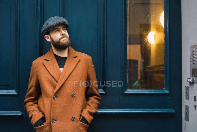 Retrato de homem pensativo vestindo casaco e boné posando na porta na rua da cidade . — Fotografia de Stock