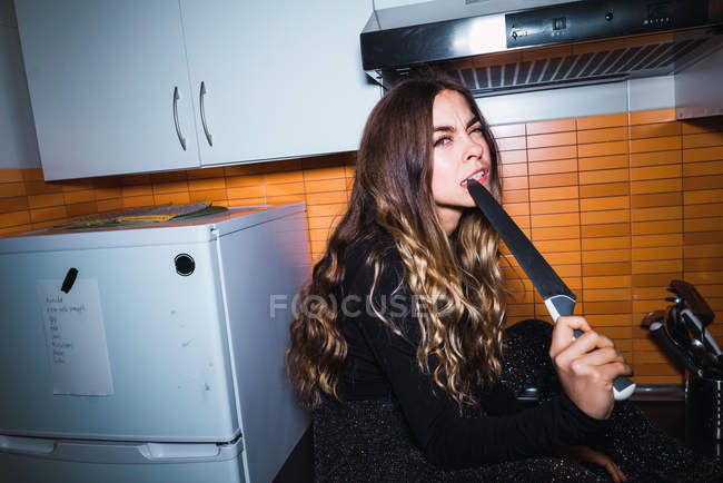 Mujer joven expresiva sentada en la mesa de la cocina y posando con cuchillo . - foto de stock
