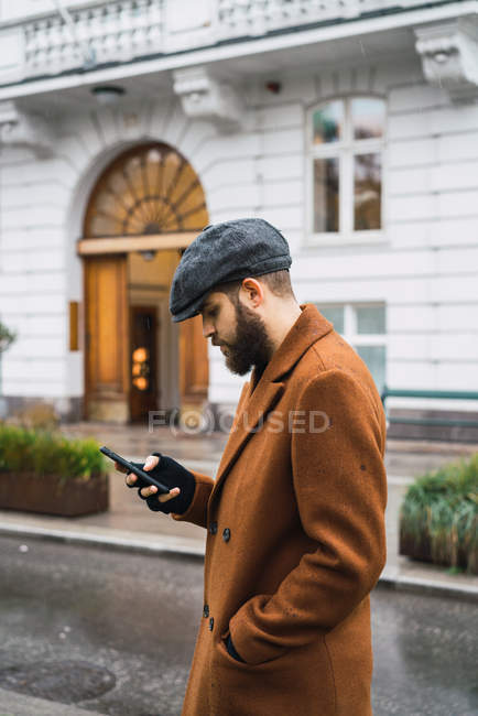 Vista laterale dell'uomo barbuto che cammina per strada e utilizza lo smartphone — Foto stock
