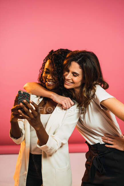 Cheerful women friends posing for selfie with smartphone on pink background. — Stock Photo