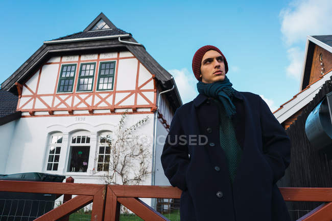 Junger Mann in warmer Kleidung steht vor traditionellem Dorfhaus an der Straße. — Stockfoto
