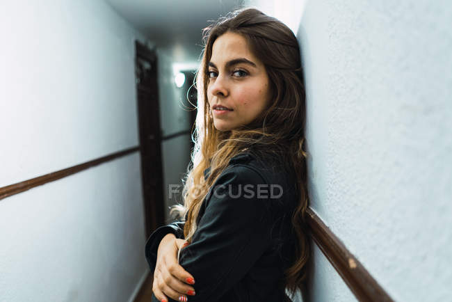 Mulher alegre inclinada na parede e olhando para a câmera no corredor do condomínio — Fotografia de Stock