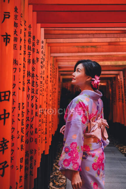 Joven bonita mujer asiática en ropa tradicional de pie en postes rojos con jeroglíficos. - foto de stock