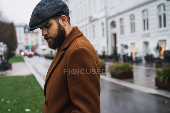 Vista lateral do homem barbudo em boné vintage e casaco posando na cena da rua — Fotografia de Stock