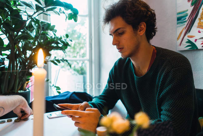 Vista lateral del joven sentado en la mesa del restaurante y el teléfono inteligente de navegación - foto de stock