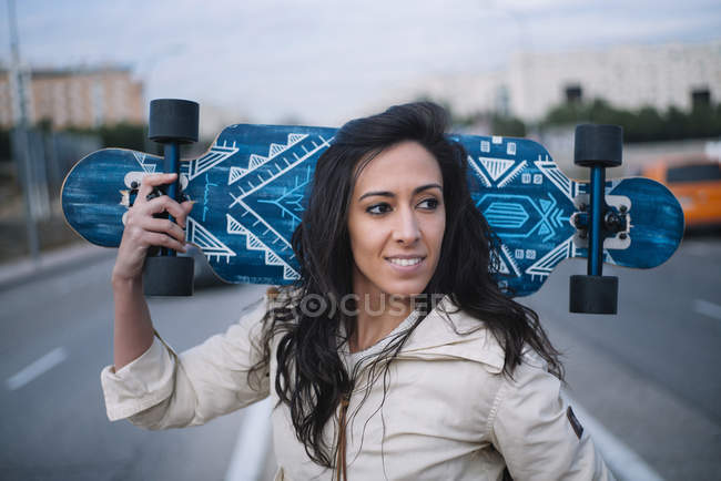 Portrait of brunette girl posing with longboard in street and looking aside — Stock Photo