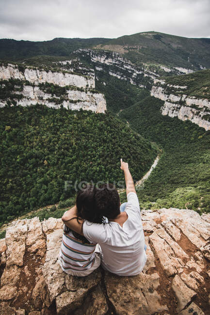 Hombre irreconocible sentado en el acantilado, abrazando a su novia y apuntando a pintorescas colinas. - foto de stock