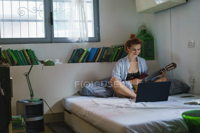 Redhead woman with guitar browsing laptop on bed — Stock Photo