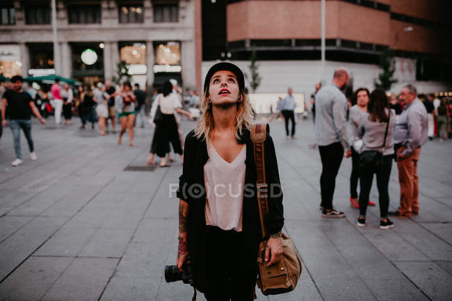 Femme avec caméra debout sur la place et levant les yeux — Photo de stock