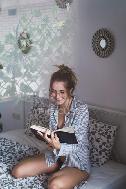 Улыбающаяся женщина в очках читает книгу на кровати — стоковое фото