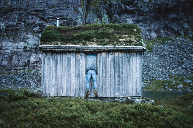 Rückansicht eines Mannes, der aus dem Fenster einer kleinen alten Hütte in den felsigen Bergen ragt. — Stockfoto