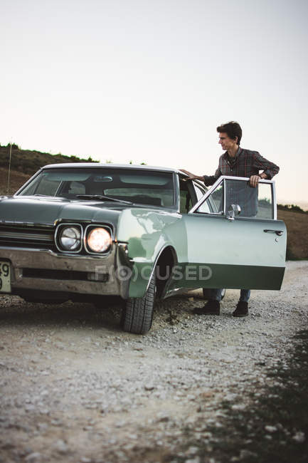 Young man getting in retro car at rural road — Stock Photo