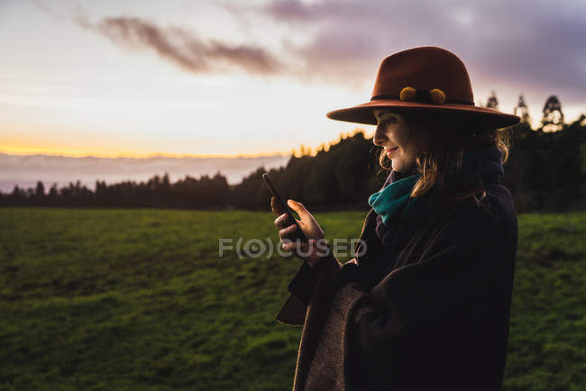 Mulher sorridente no smartphone de navegação de chapéu em campos frios verdes no crepúsculo — Fotografia de Stock