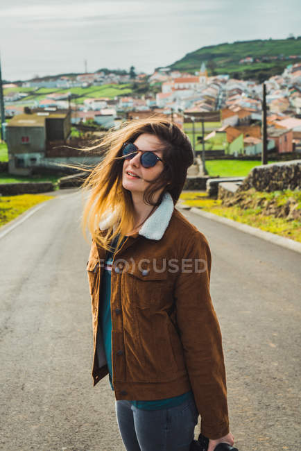 Стильная молодая женщина в солнечных очках, стоящая на дороге на фоне прибрежного города . — стоковое фото