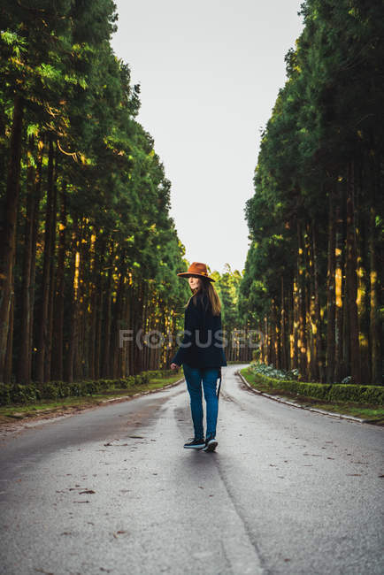 Visão traseira da jovem caminhando na estrada rural e olhando sobre o ombro na câmera — Fotografia de Stock